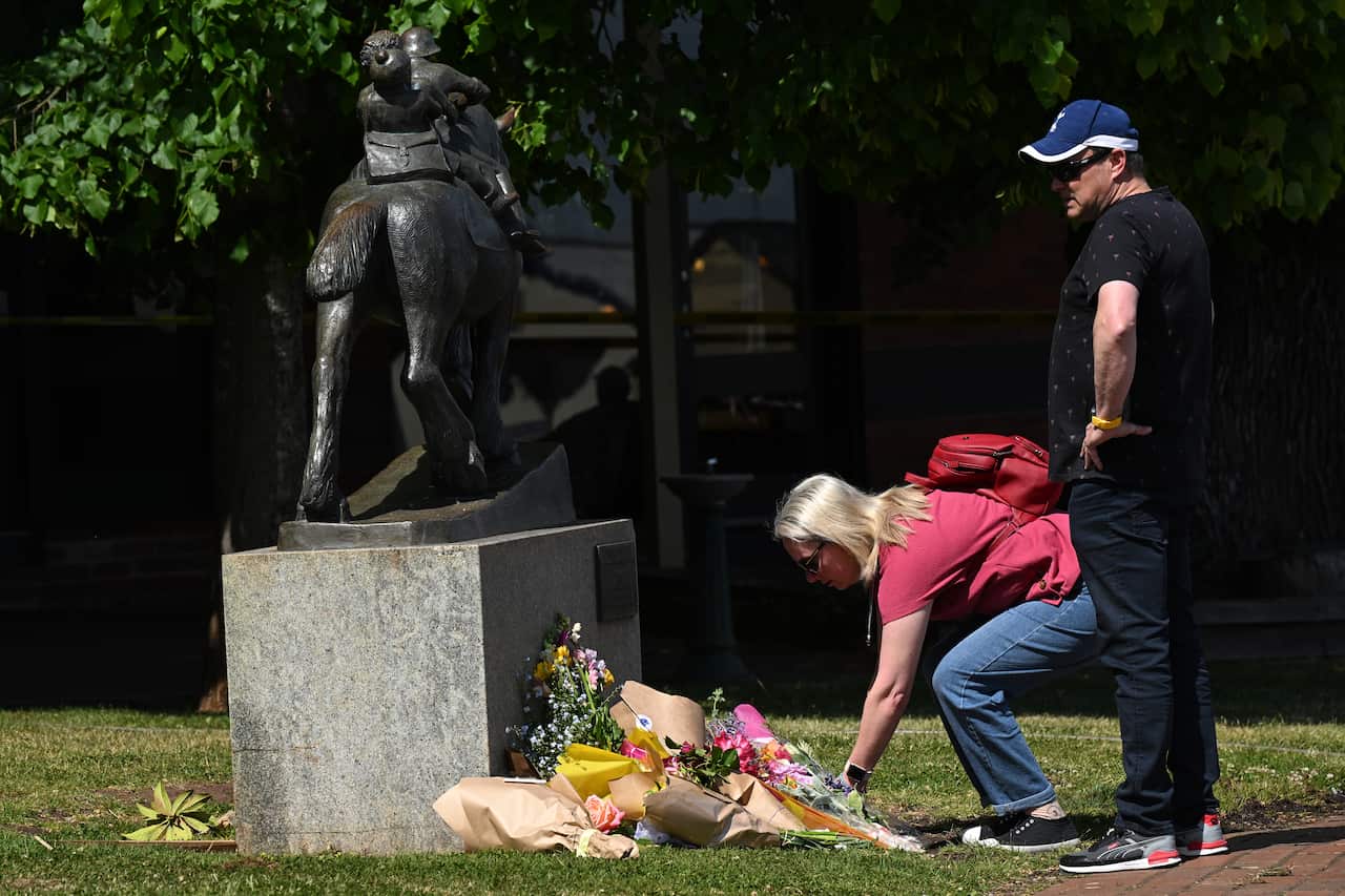 A woman leaves flowers at a makeshift memorial while a man in a baseball hat watches on.