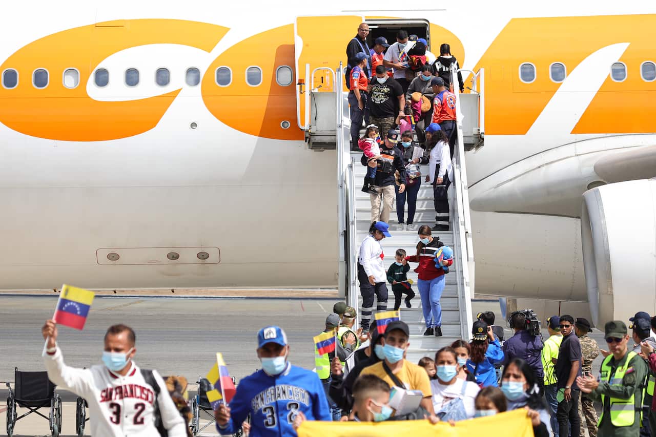 Passengers disembark from a plane while holding Venezuelan flags, with a crowd welcoming them at the airport.