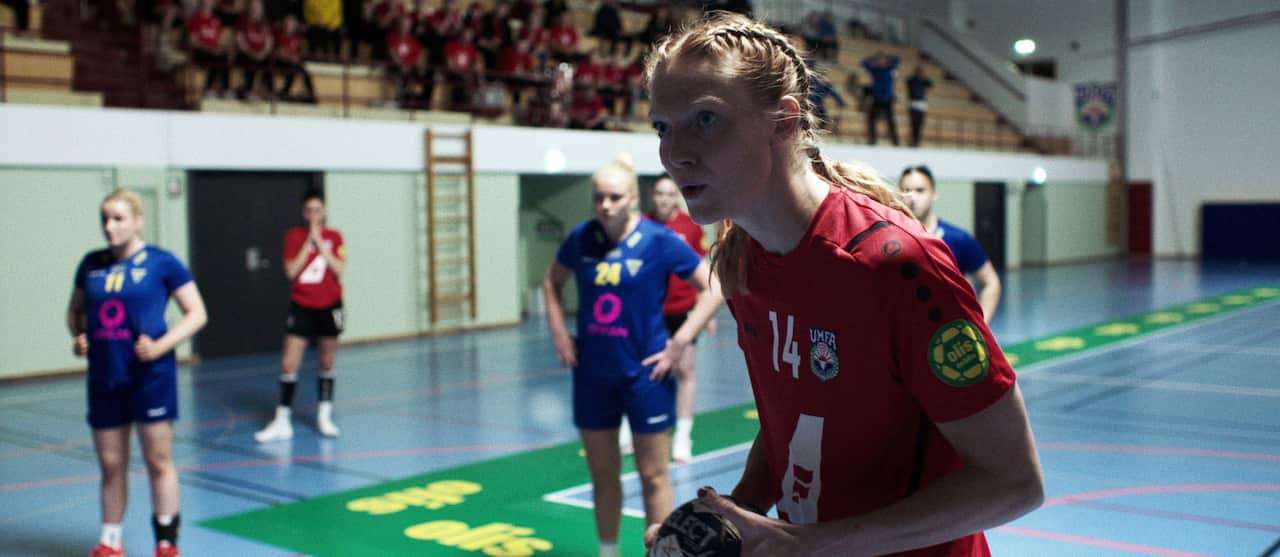 A young woman in a red shirt stands on a court, holding a ball. Other women stand behind her, some in the same team shirts and some in a blue uniform. 