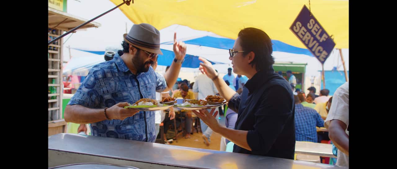 Chef Luke Nguyen (right) with a plate in one hand and pointing with another man underneath a tarpaulin.