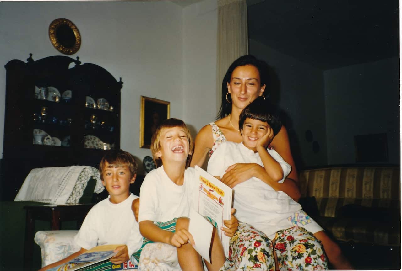 Woman in a floral jumpsuit holds a young boy on her lap with two other boys in white t-shirts by her side holding children's books