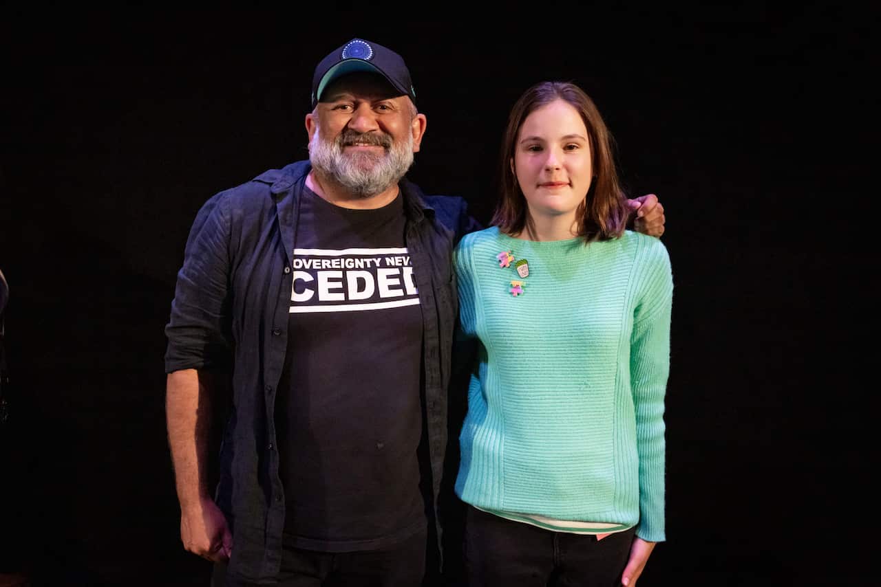 A middle aged Gomeroi man has his arm around the shoulders of a young white girl. They smile at the camera. 