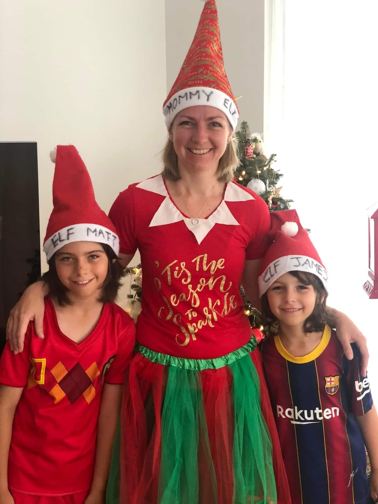 A woman dressed in Christmas dress wears a Santa hat with the words 'Mommy Elf' on it, with her arms around her children.