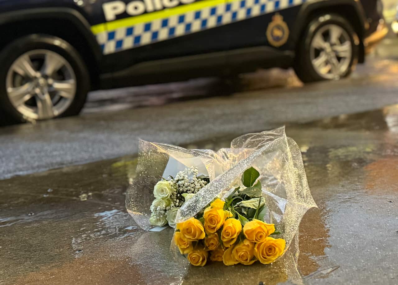 Two bouquets of flowers, wrapped in plastic, lying on a wet ground. A police vehicle is in the background.
