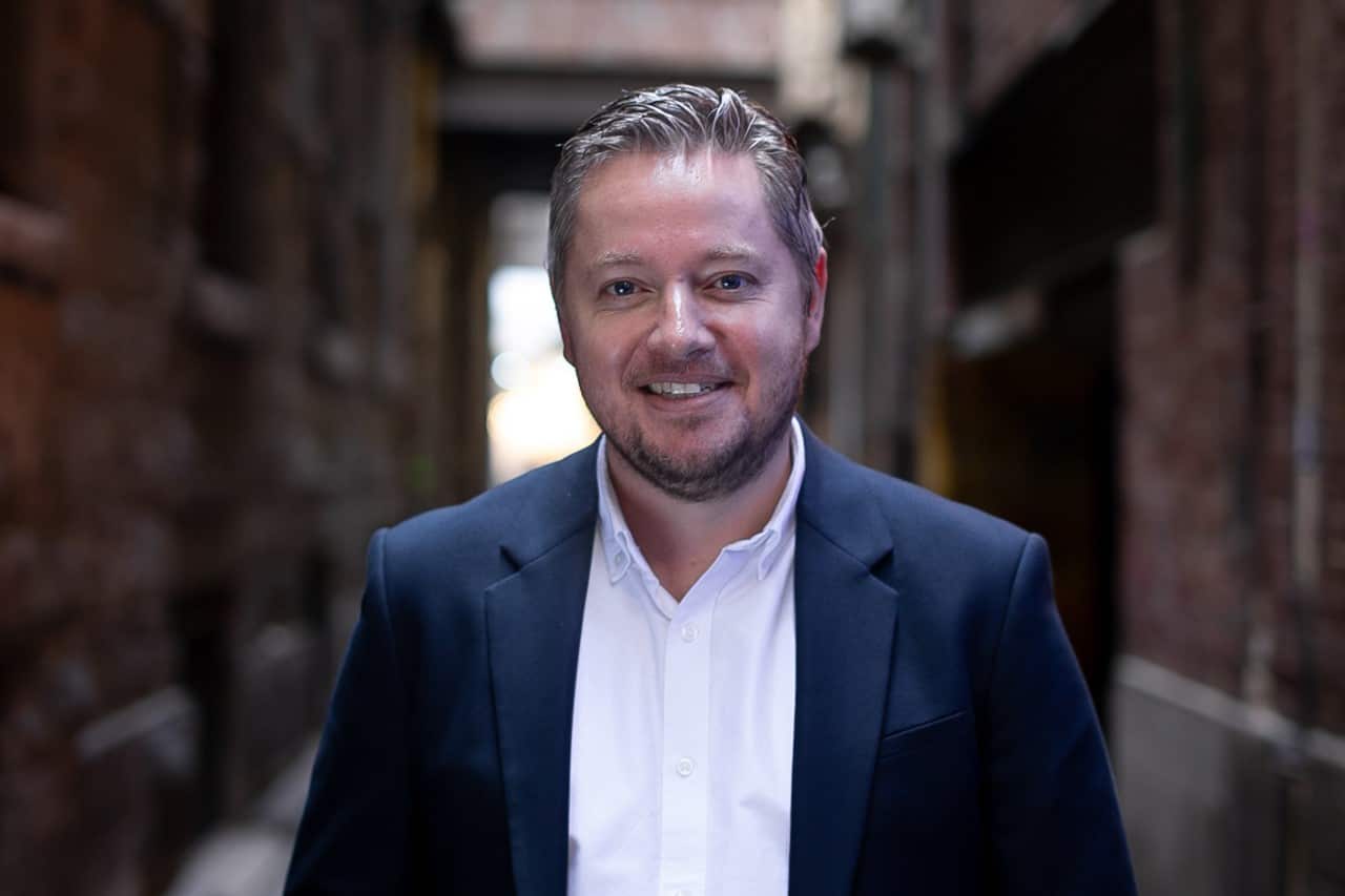 A man in a blue coat smiles as he stands in a laneway.