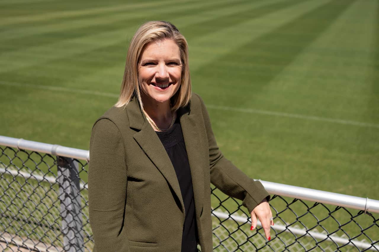 A middle-aged white woman smiling. She's leaning against a fence, a football pitch is behind her.