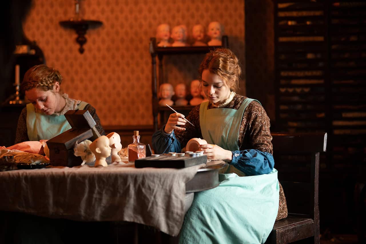 Two young women site at a table, painting dolls faces. 