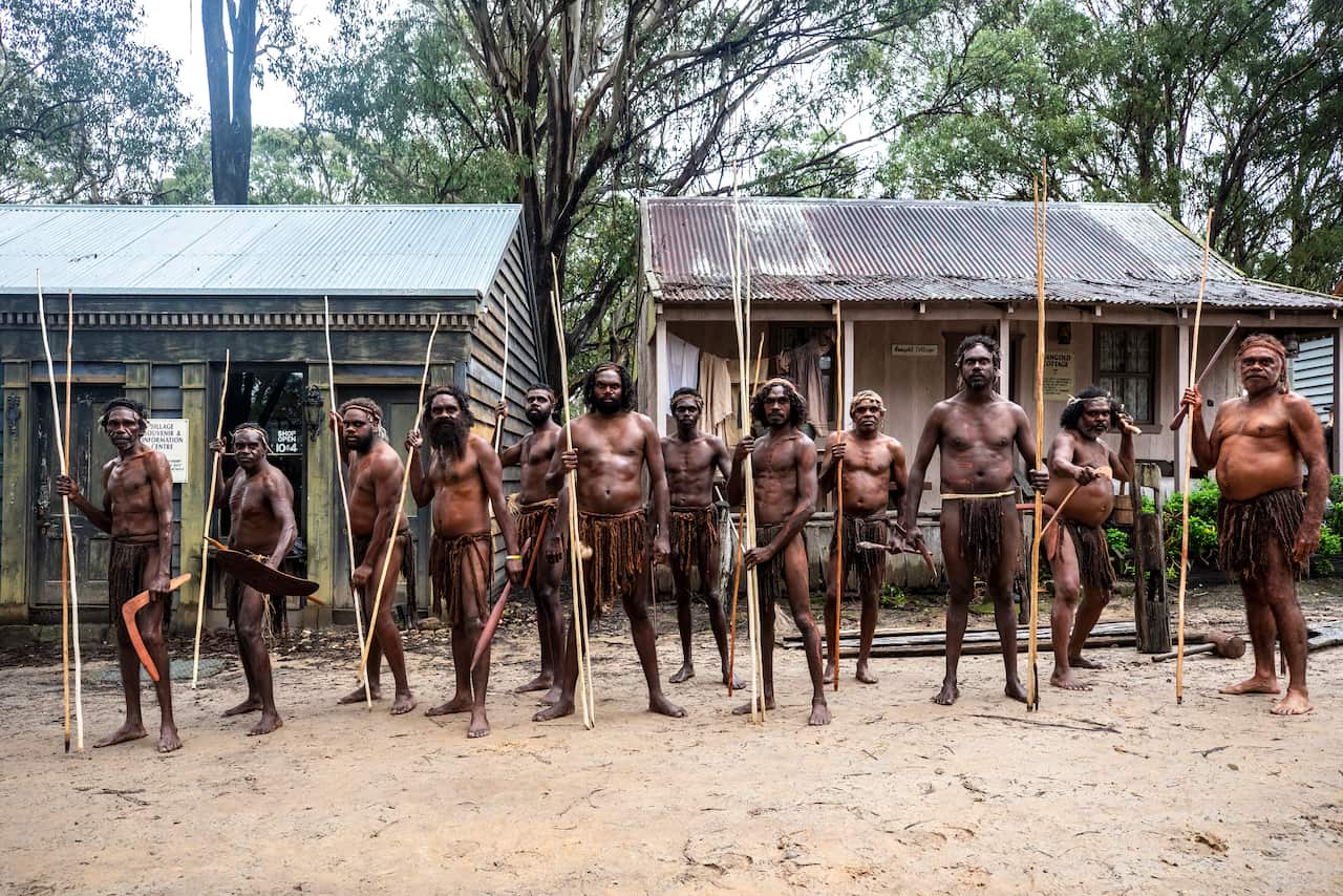 Pemulwuy and a band of warriors stand firm and hold spears amongst the shacks of a colonial settlement.