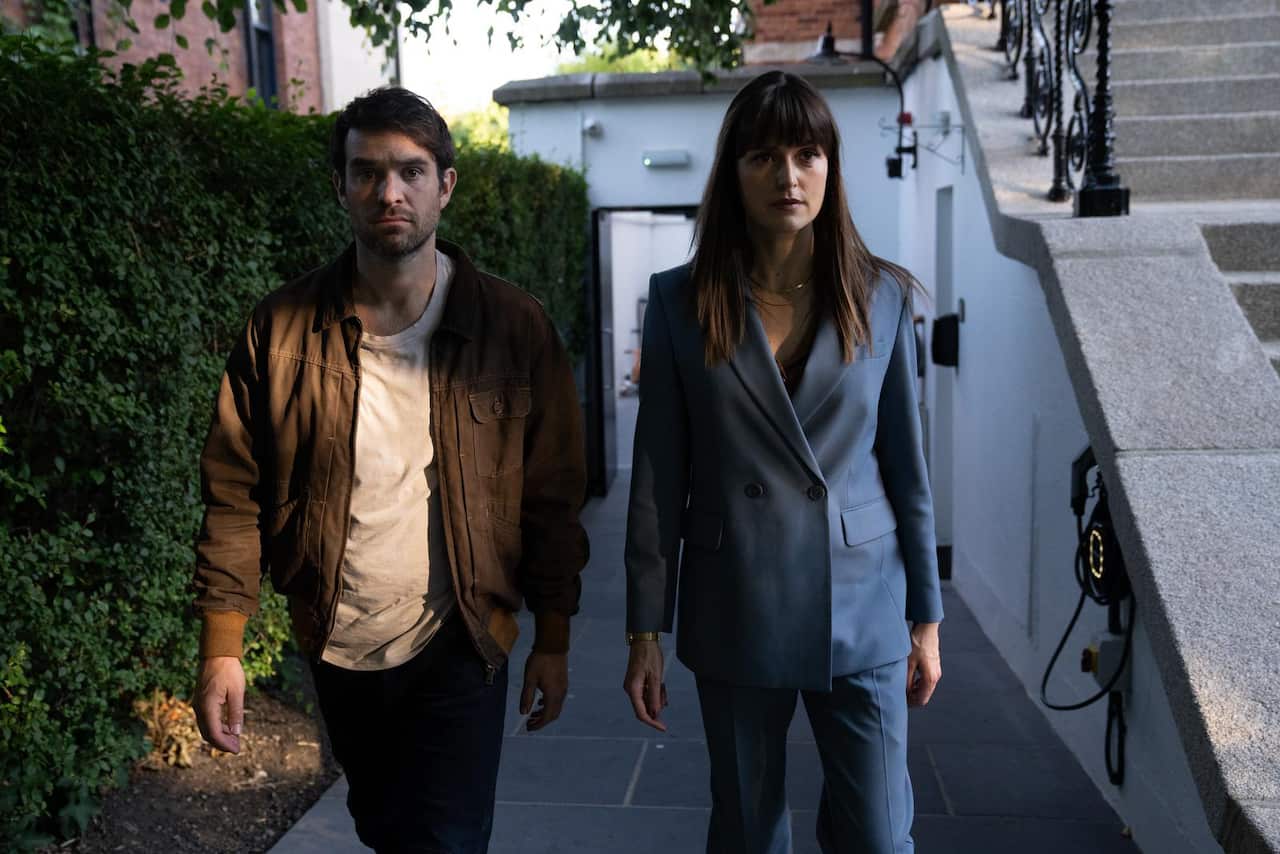 A woman in a suit and a man in a brown jacket walk side by side on a bath beside a house. 