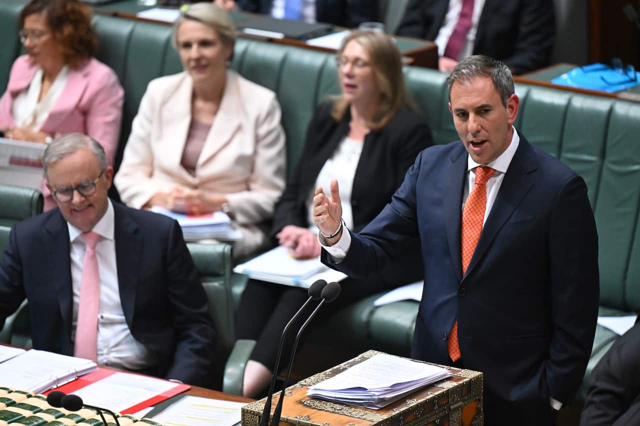 A man in a suit is standing as he speaks into microphones with one hand raised. There are several people in formal attires sitting in rows behind him.