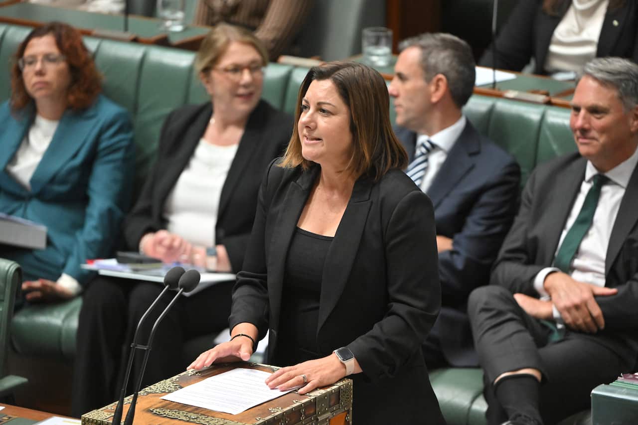 A woman in a black top and blazer is speaking into the microphones in the parliament.