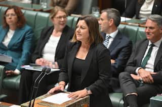 A woman in a black top and blazer is speaking into the microphones in the parliament.