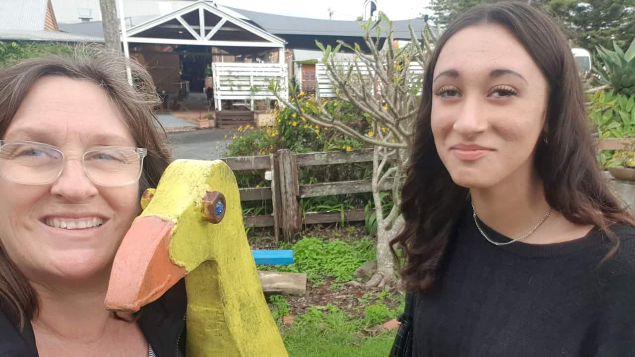 a middle aged woman with clear frame glasses and shoulder length brown hair holds a toy wooden duck and is standing with a smiling teenage girl in a black jumper. They are in front of a rural home