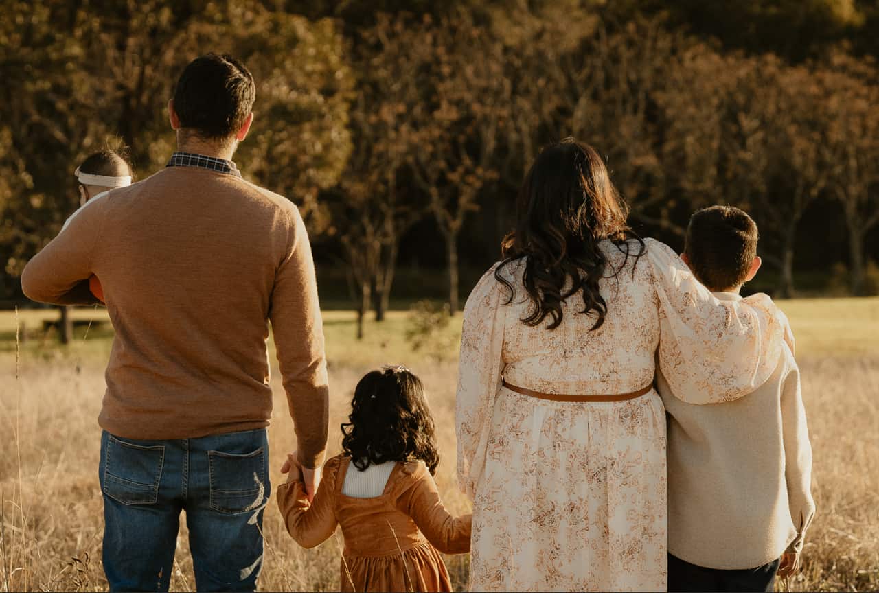 A mum, dad and their three young children stand with their backs to the camera in a wheat field.