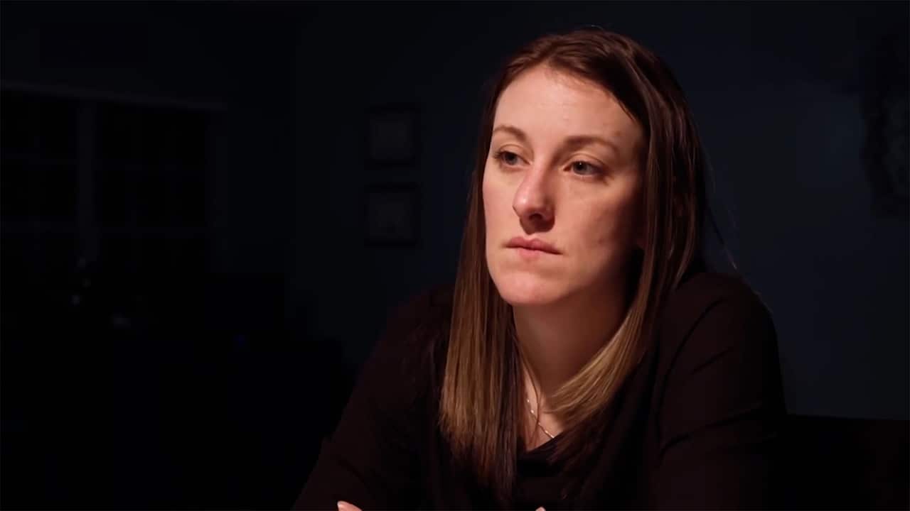 Nathalie Collins stares into the distance. She wears a black top and is surrounded by darkness, with shadows casting over her face.