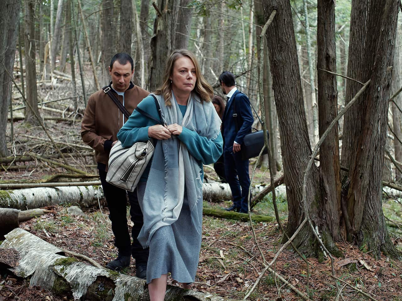 A woman in a blue dress, clutching a bag, walks through a wood. Two men can be seen behind her.