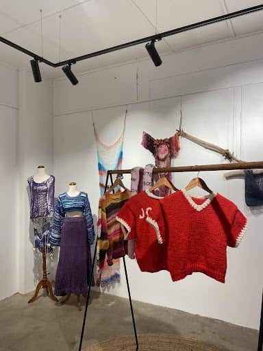 Red and multicoloured knitted tops hung from a hanger with dresses and tops displayed in the background in a hall.