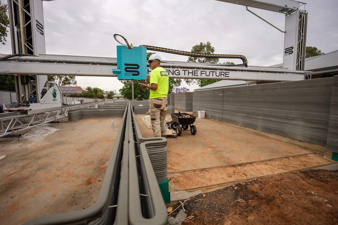 A worker on a construction site, working with a large 3D printing machine, printing the walls of a house with concrete, one layer at a time.