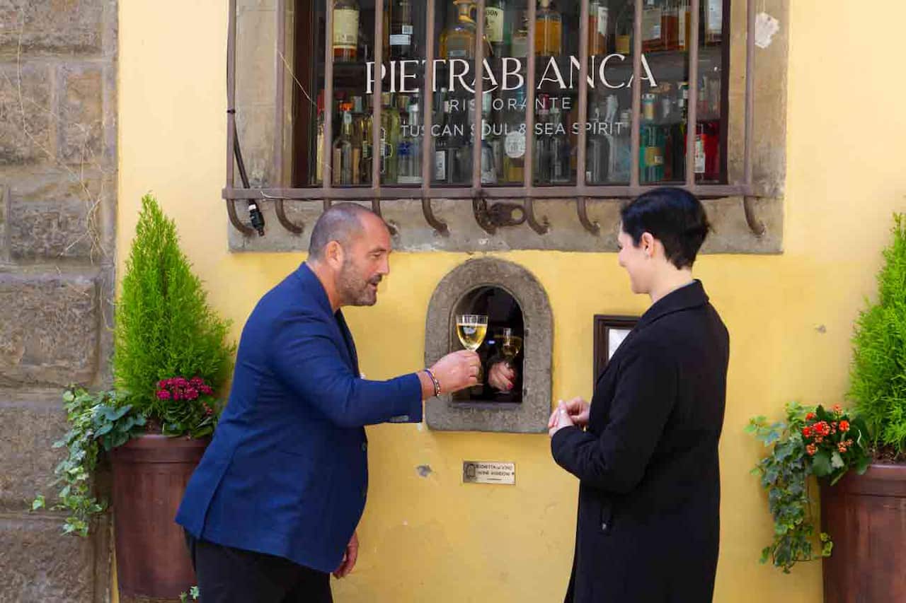 A man and woman stand in front of a yellow wall. Between them, in tiny door in the wall, at shoulder height, a hand holds out a glass of wine. 