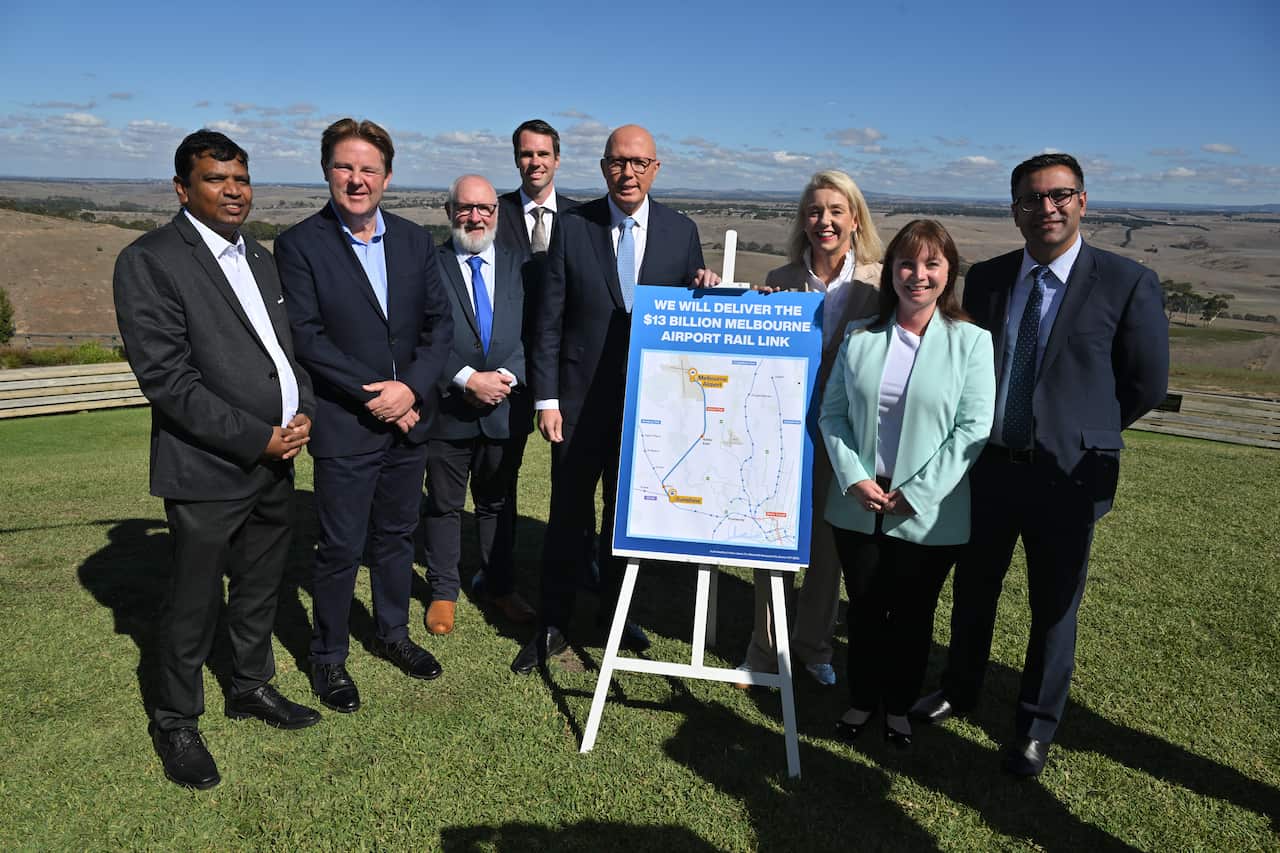A group of people are smiling as they stand behind a signboard in the grass. 