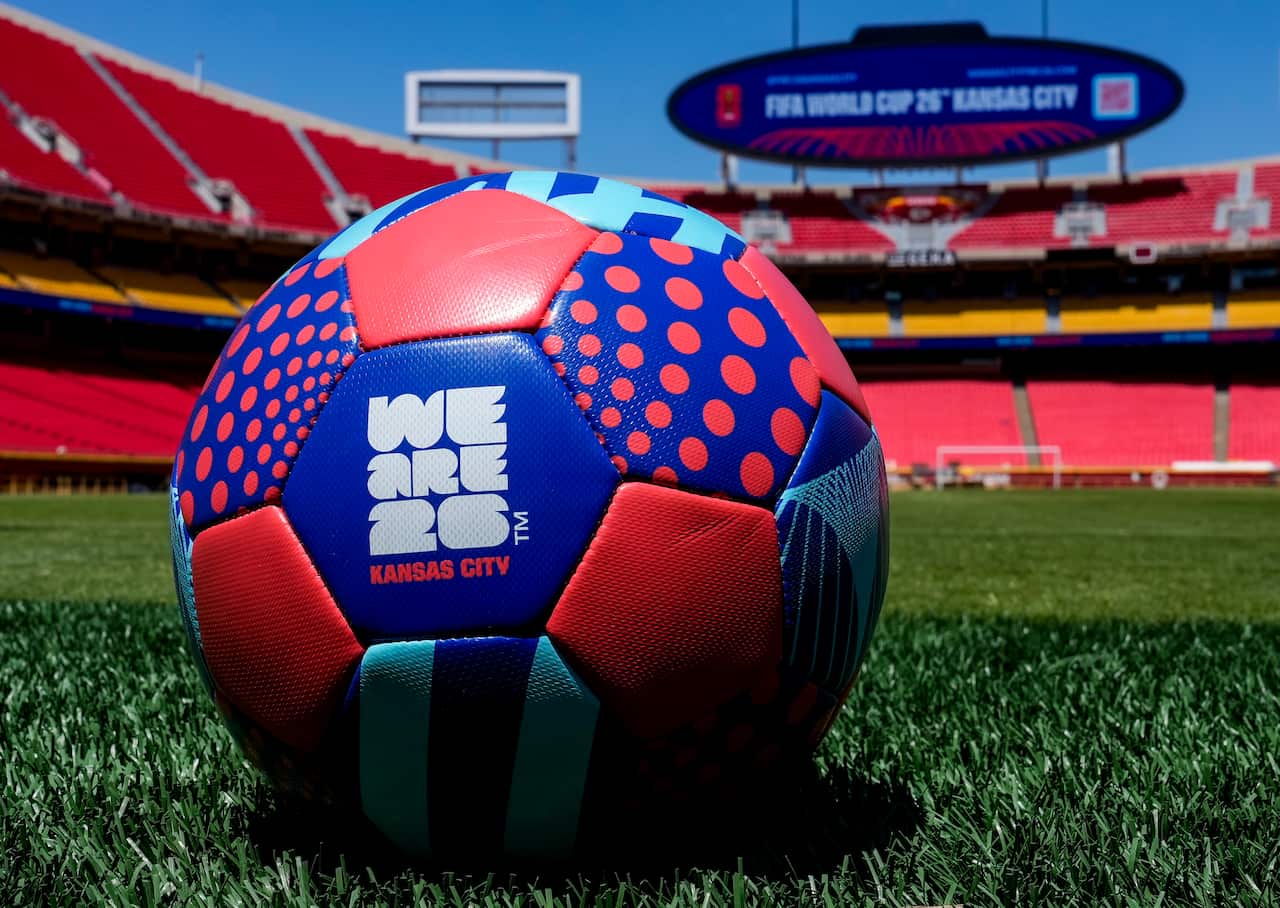 A multi-coloured football on the grass in a large stadium. One of the ball's panels has We Are 26 in white print on it and below in smaller red print, Kansas City