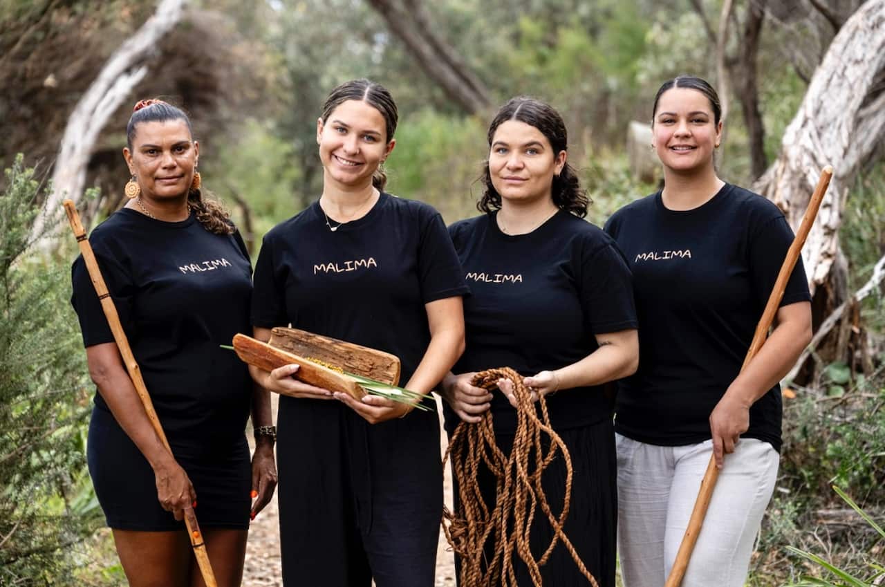 A group of First Nations women wear black t-shirts which read 'Malima' while standing in bushland. They hold traditional items including spears and woven ropes.