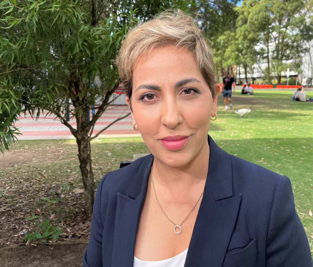 A woman in a blue jacket sits in a grassed area near a small tree.