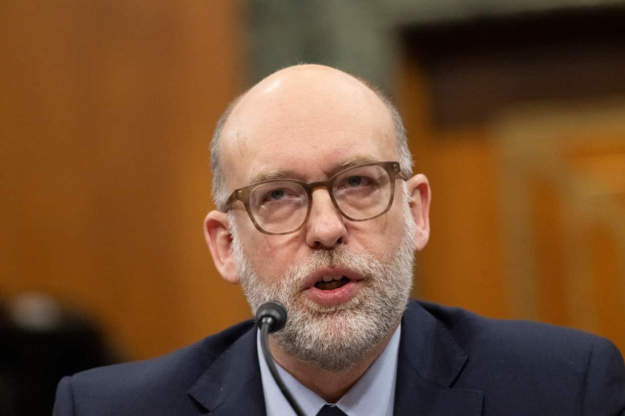 A man wearing a suit and glasses speaks at a lectern. 