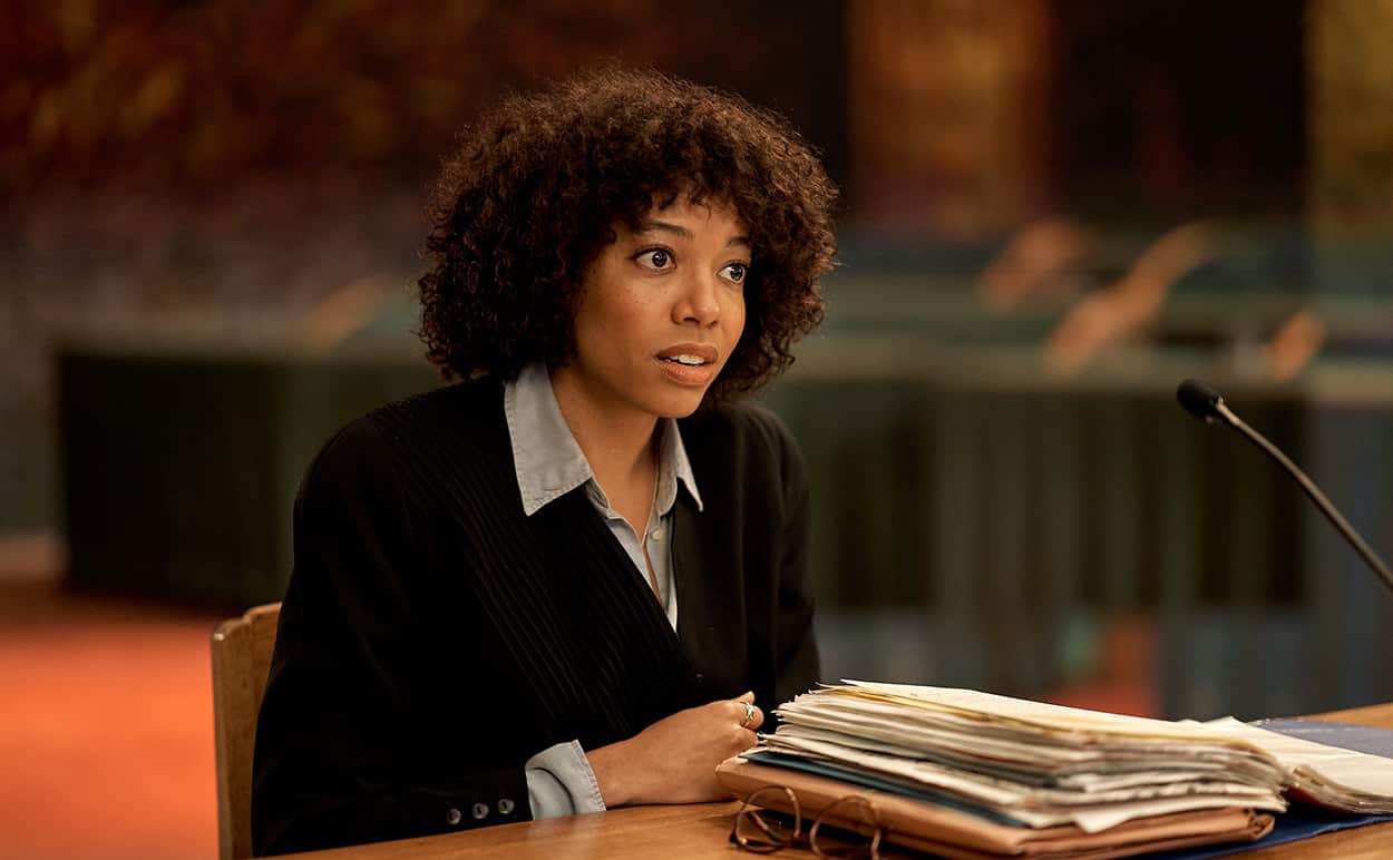 Woman with curly hair in dark suit sits at table in front of microphone.