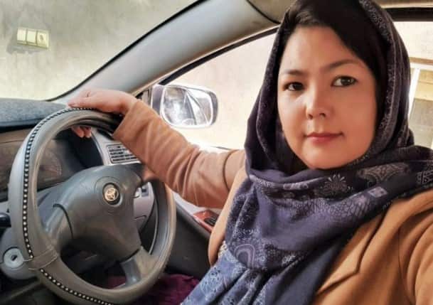 A woman in a brown jacket and headscarf sits behind the steering wheel of a car.