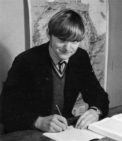A black and white photo of a smiling teenager at a desk, holding a pen. He sits in front of a map and is wearing a dark jacket, jumper and tie.