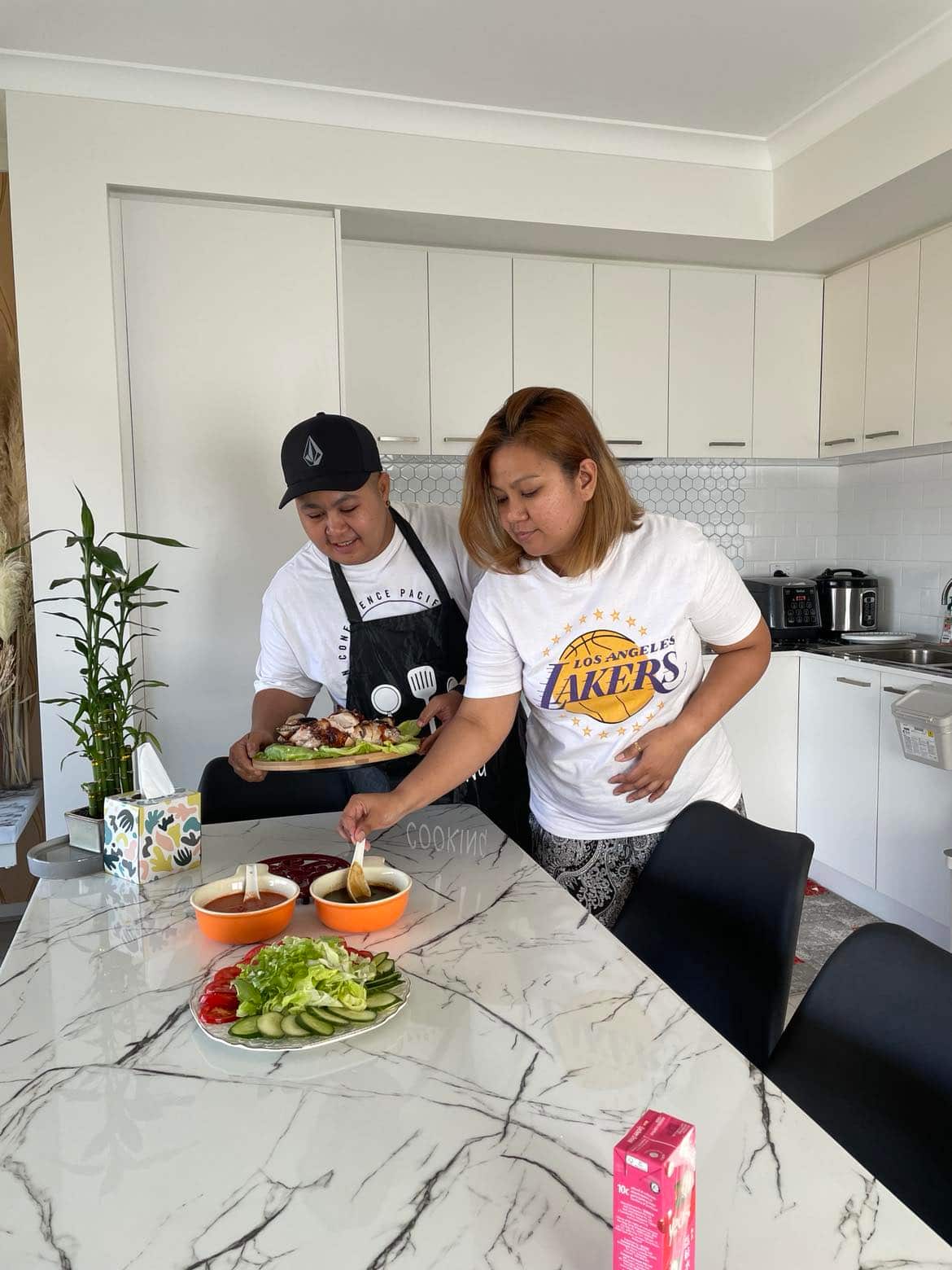 Two people in white T-shirts prepare a meal together in a kitchen.