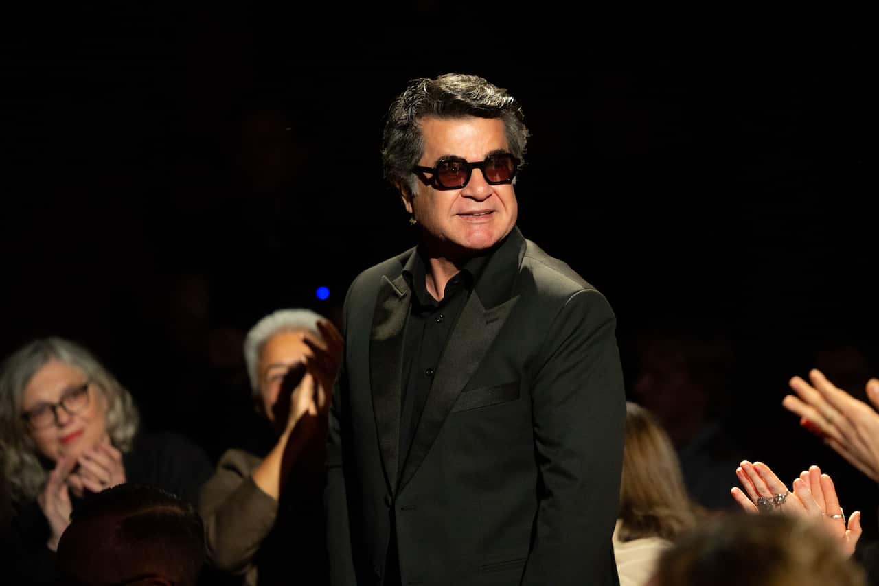 A man in a black suit and sunglasses stands and smiles as people sitting around him clap.
