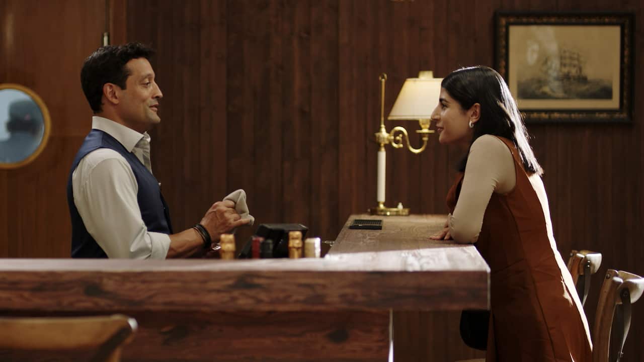 A woman leans on a wooden bar. A barman stands on the other side, smiling. 