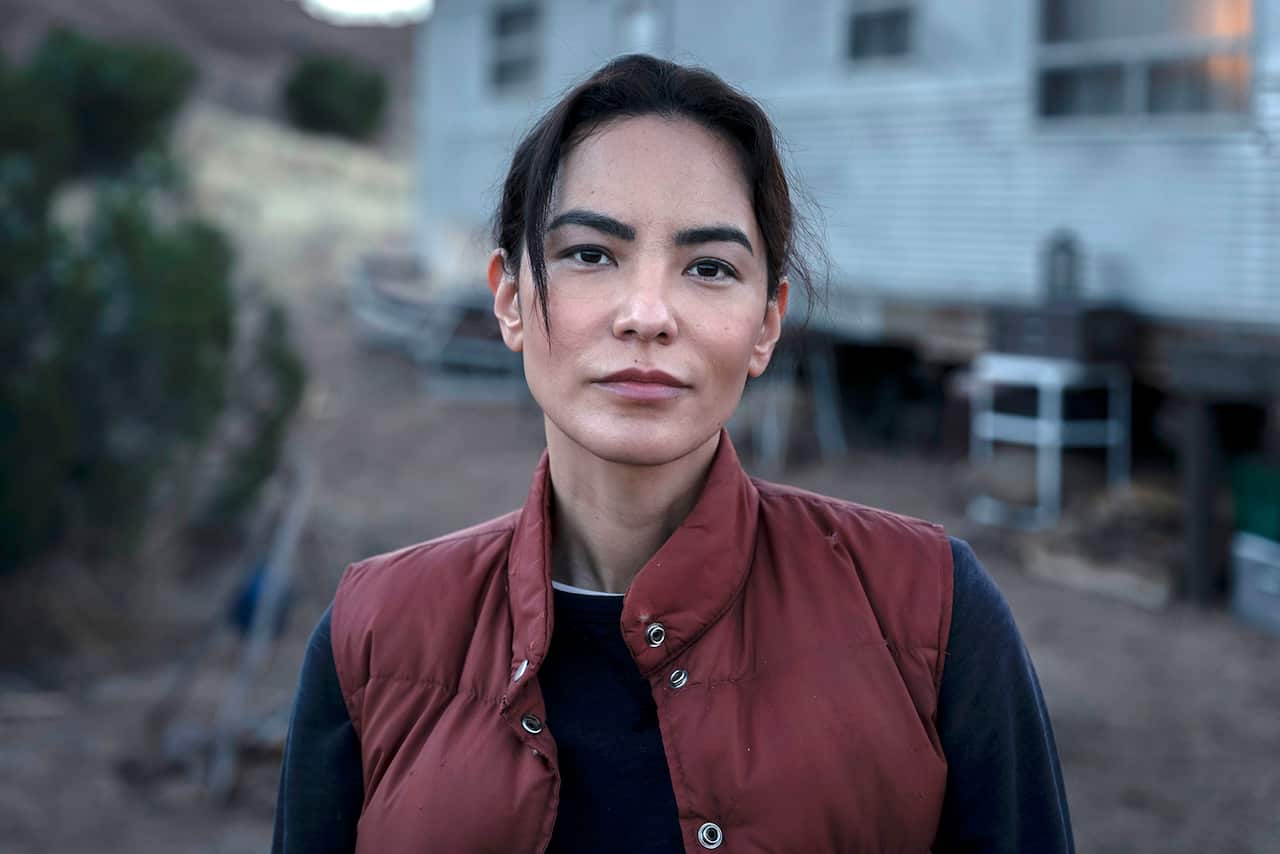 A dark-haired woman in a dark red vest and dark shirt stares staight at the camer.