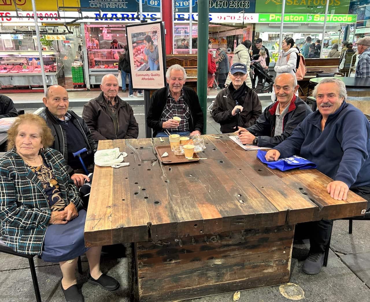 A group of older men and a woman sit at a large wooden table in a market walkway.