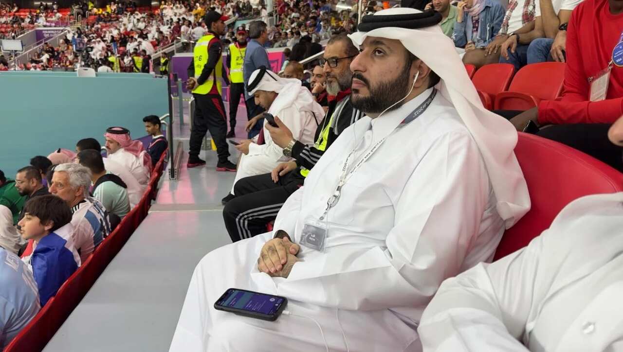 A man in a white robe in a football stadium wearing earphones connected to a mobile phone on his lap