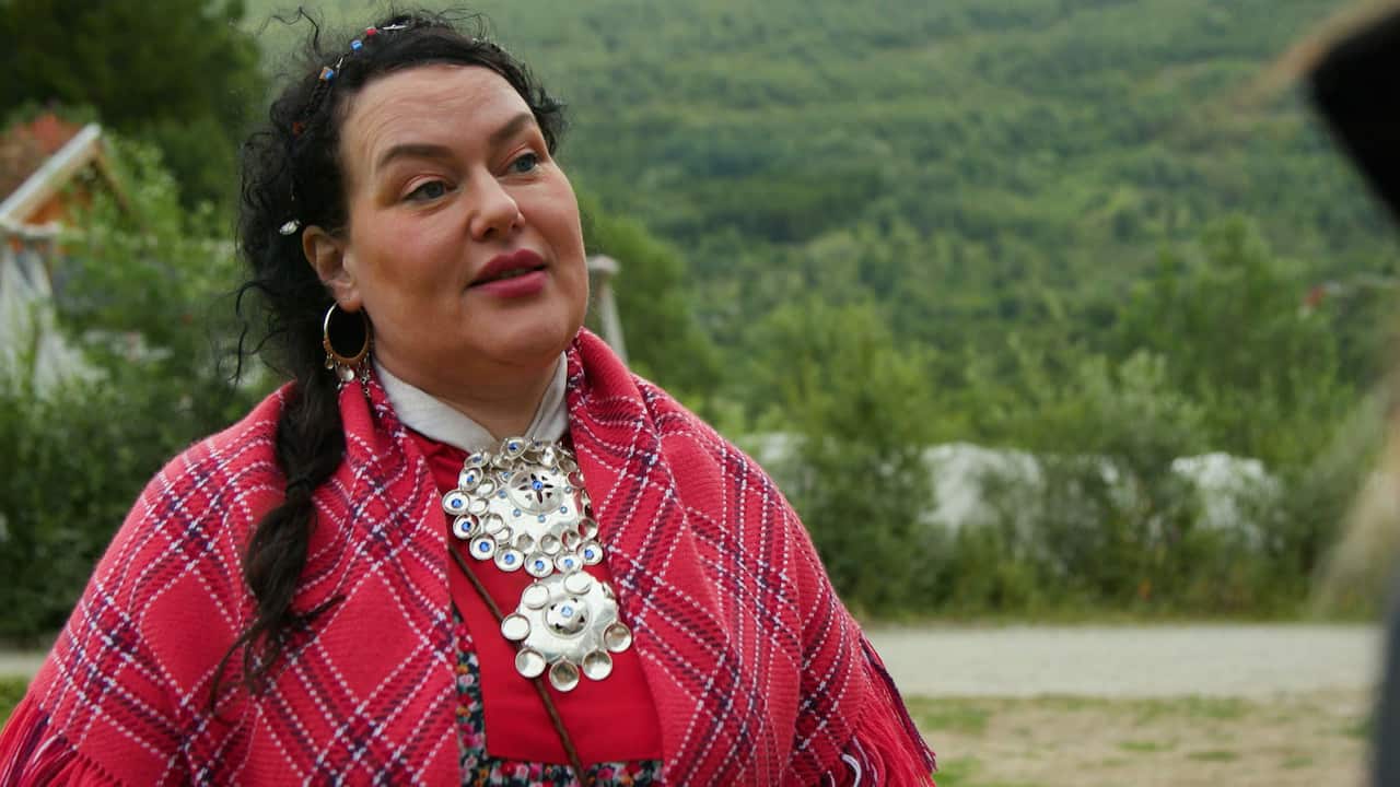 A woman with dark hair wearing a traditional outfit of the Indigenous Sami people: a red dress with a red tartan cape, and a large silver necklace. 