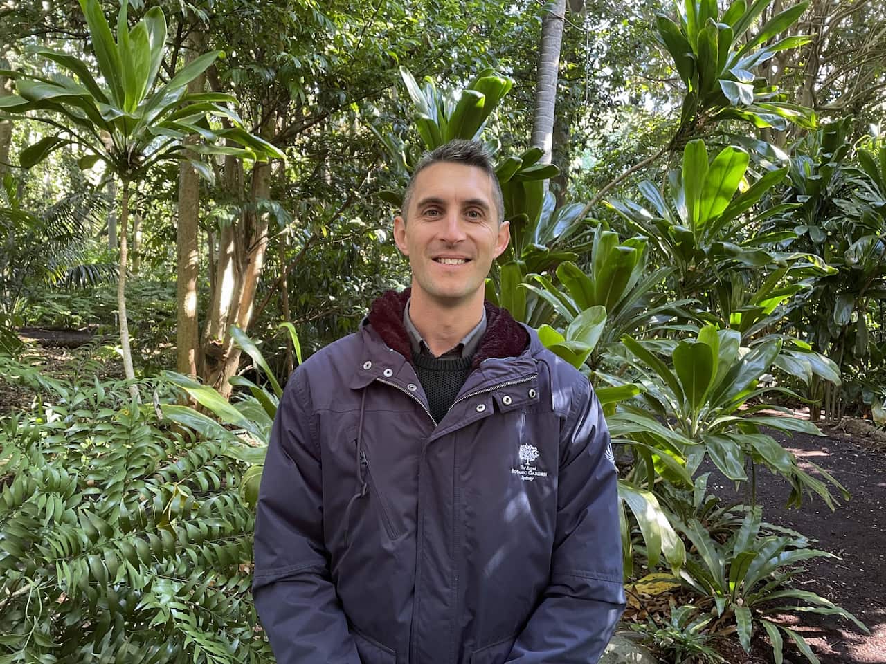 A man in a blue jacket standing outside with green plants behind him.