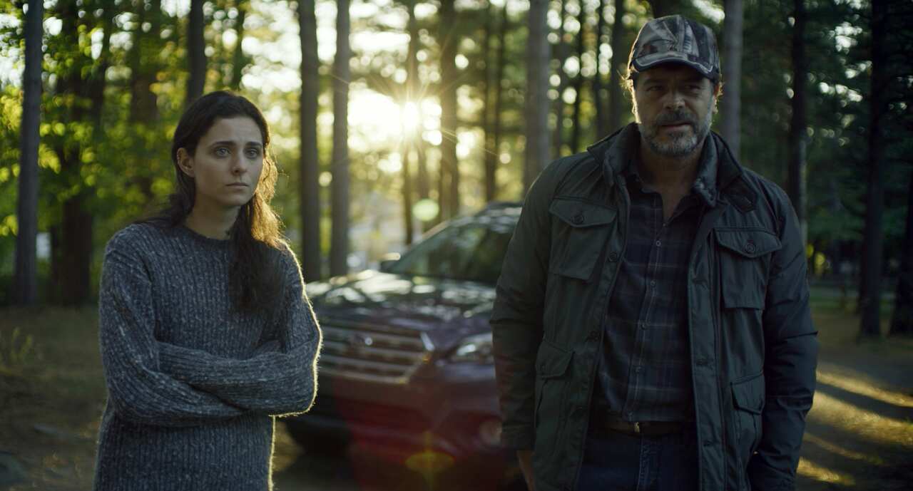 A young woman and man stand in a clearing, with a car and trees behind them. 