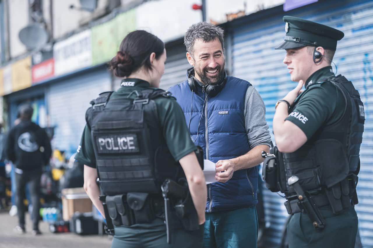 Two actors in police clothing stand talking to a man wearing headphones. 