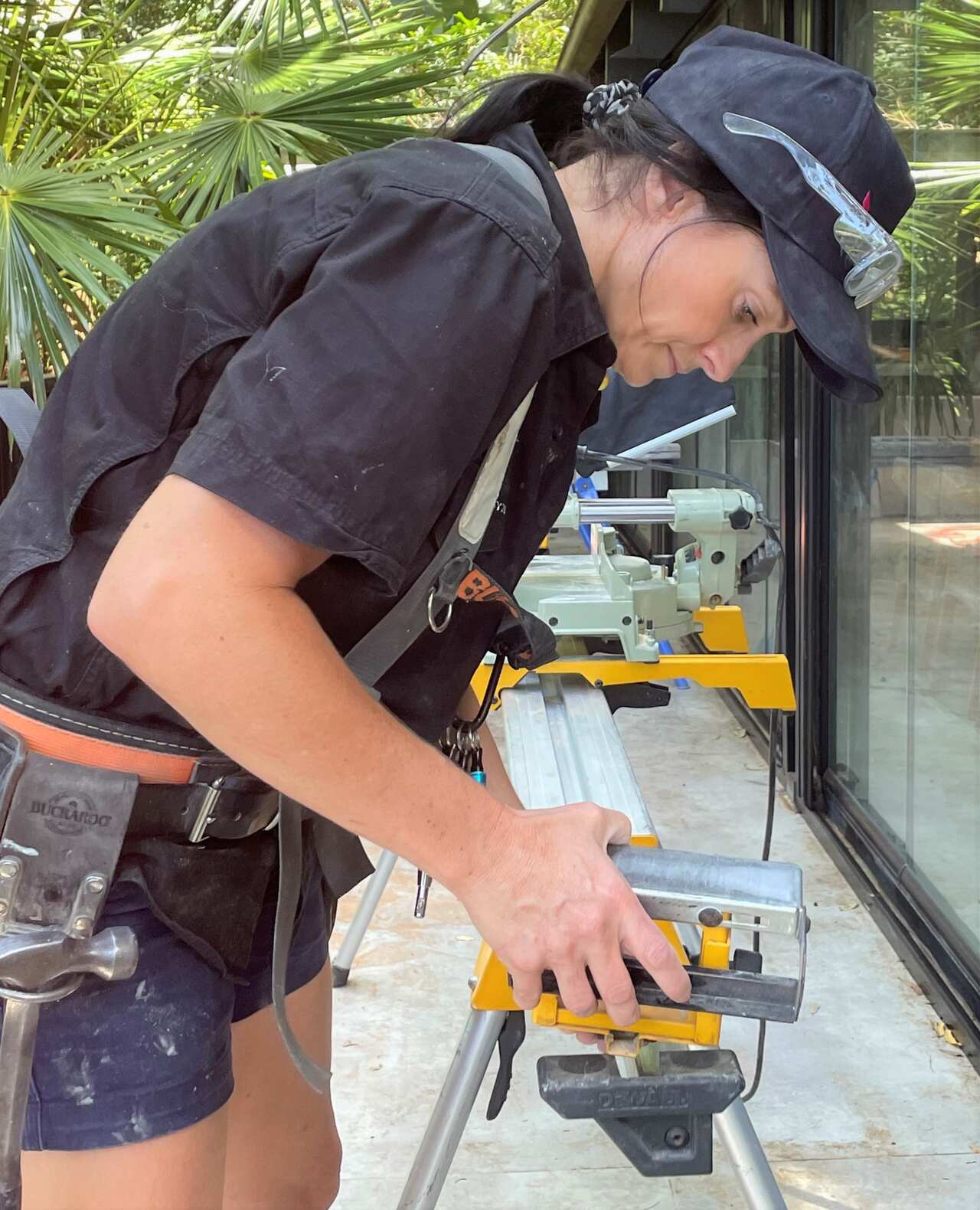 A trades woman wearing work clothes leans over a work bench using a cutting tool.