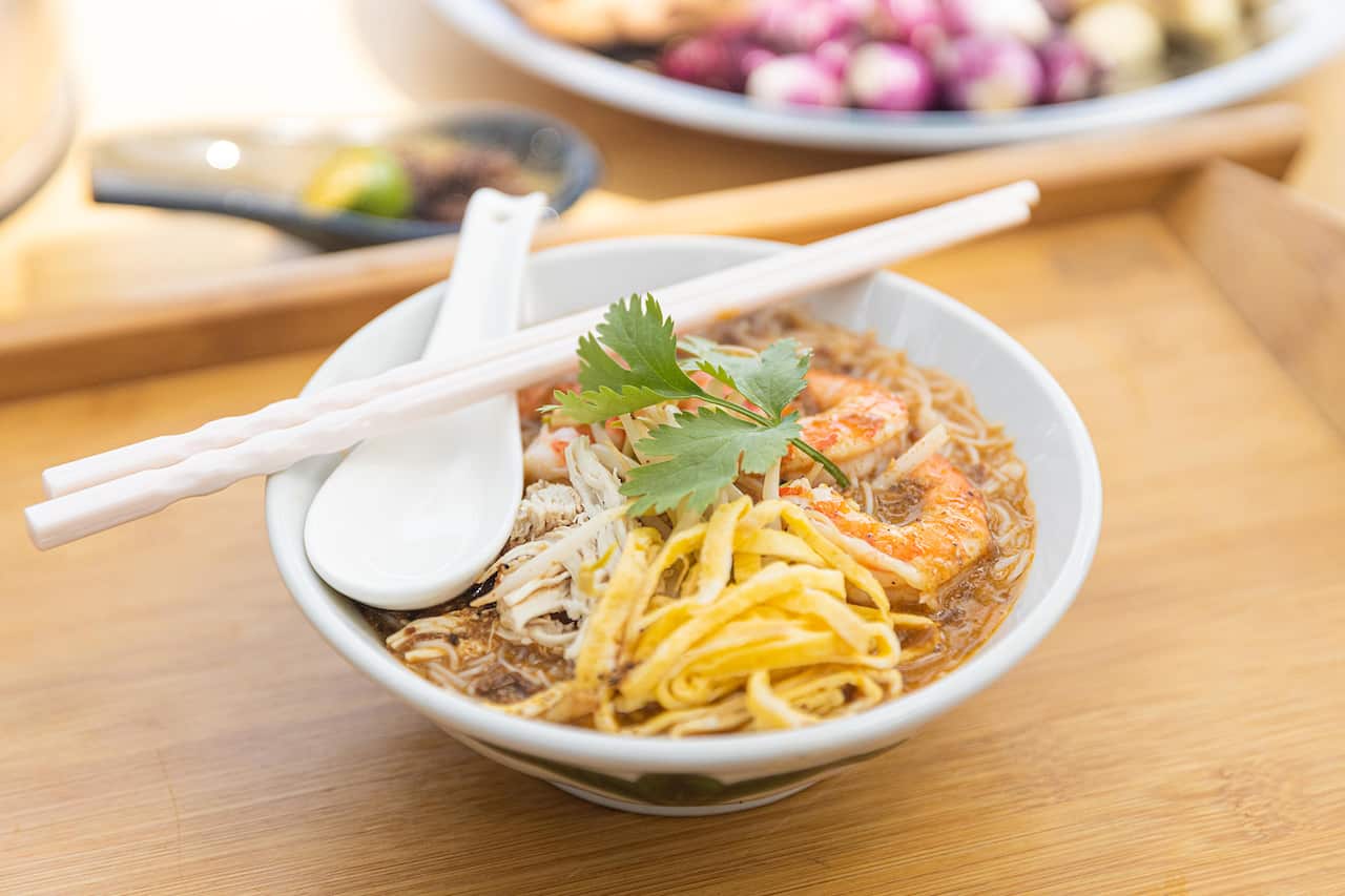 A white bowl sits on a wooden surface. It holds a laksa, with prawns visible. 