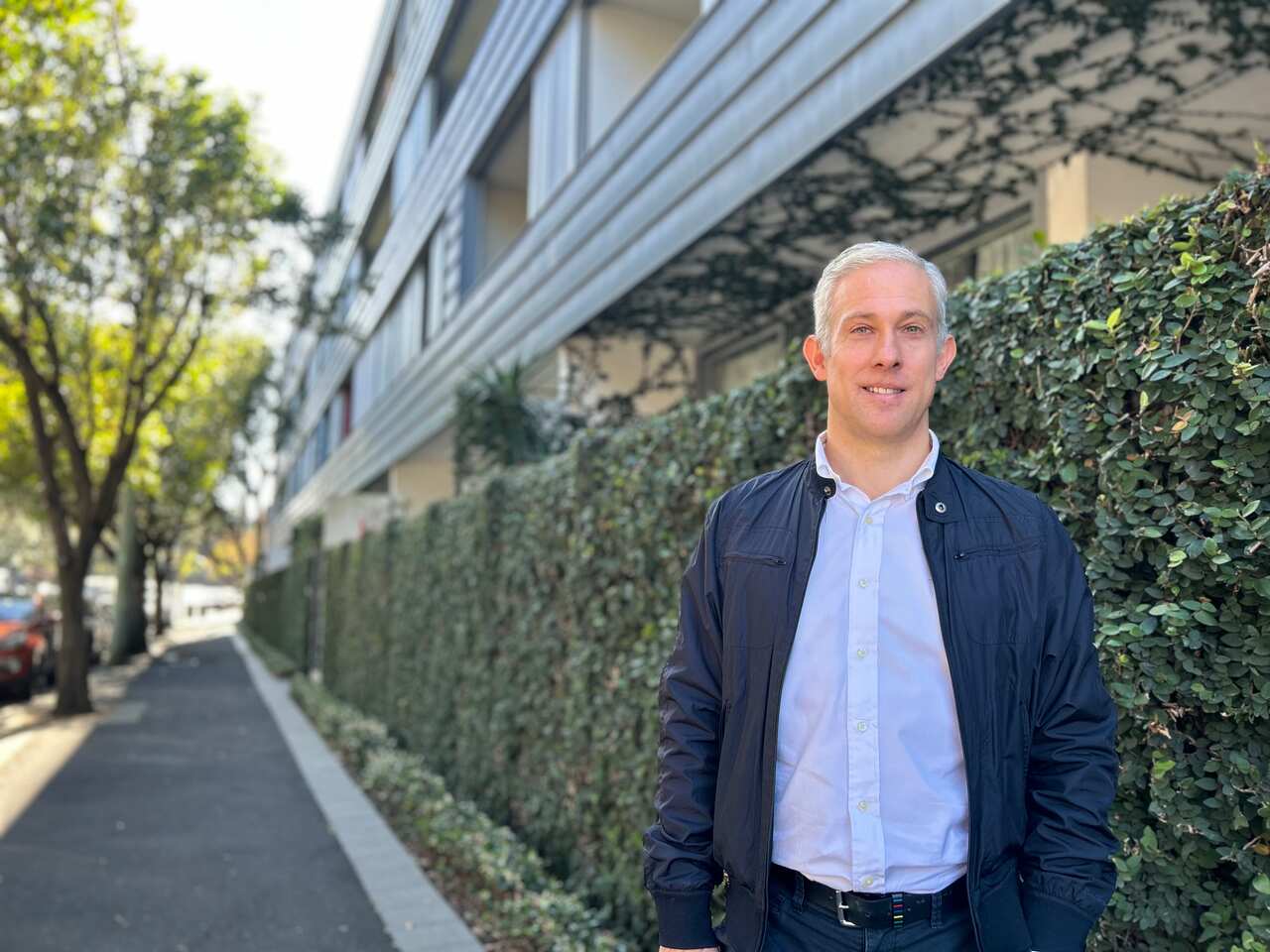 A man stands in front of an apartment building's outer wall, which is covered in foliage. 