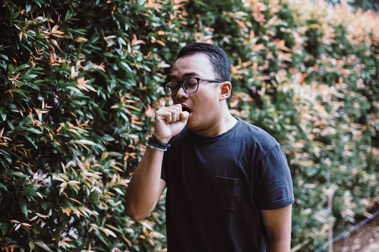 A man walking outside, holding his fist to his mouth as he coughs.