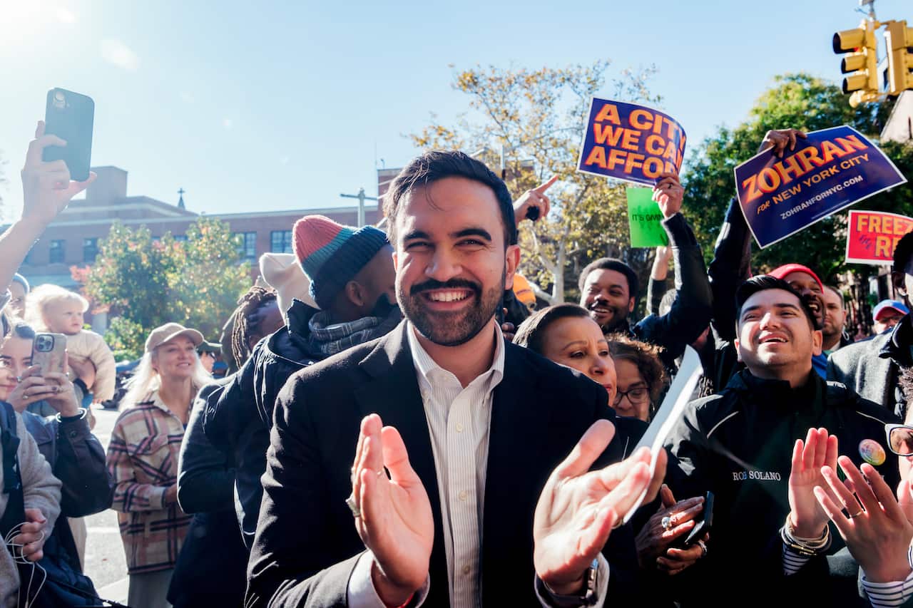 A young man wearing a dark suit and white shirt claps and smiles among a crowd of supporters. 