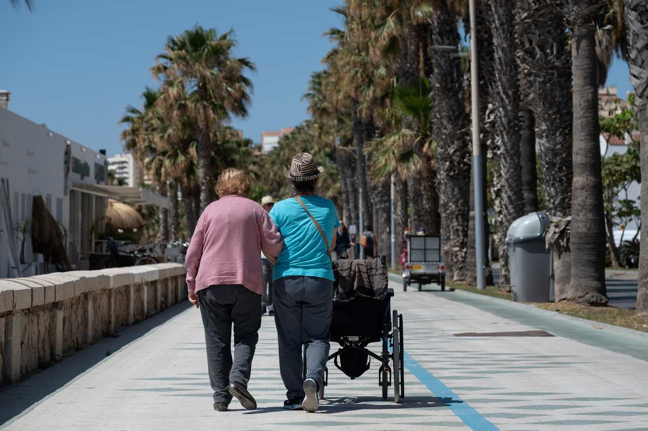 Two elderly women are walking on a path surrounded by palm trees. One of them is pushing a wheelchair.