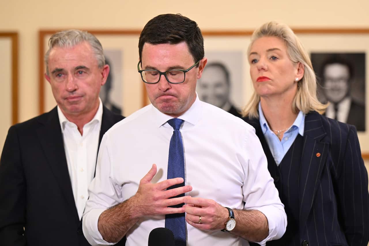 A man in a white shirt and blue tie stands with a man and a woman in dark blazers behind him.