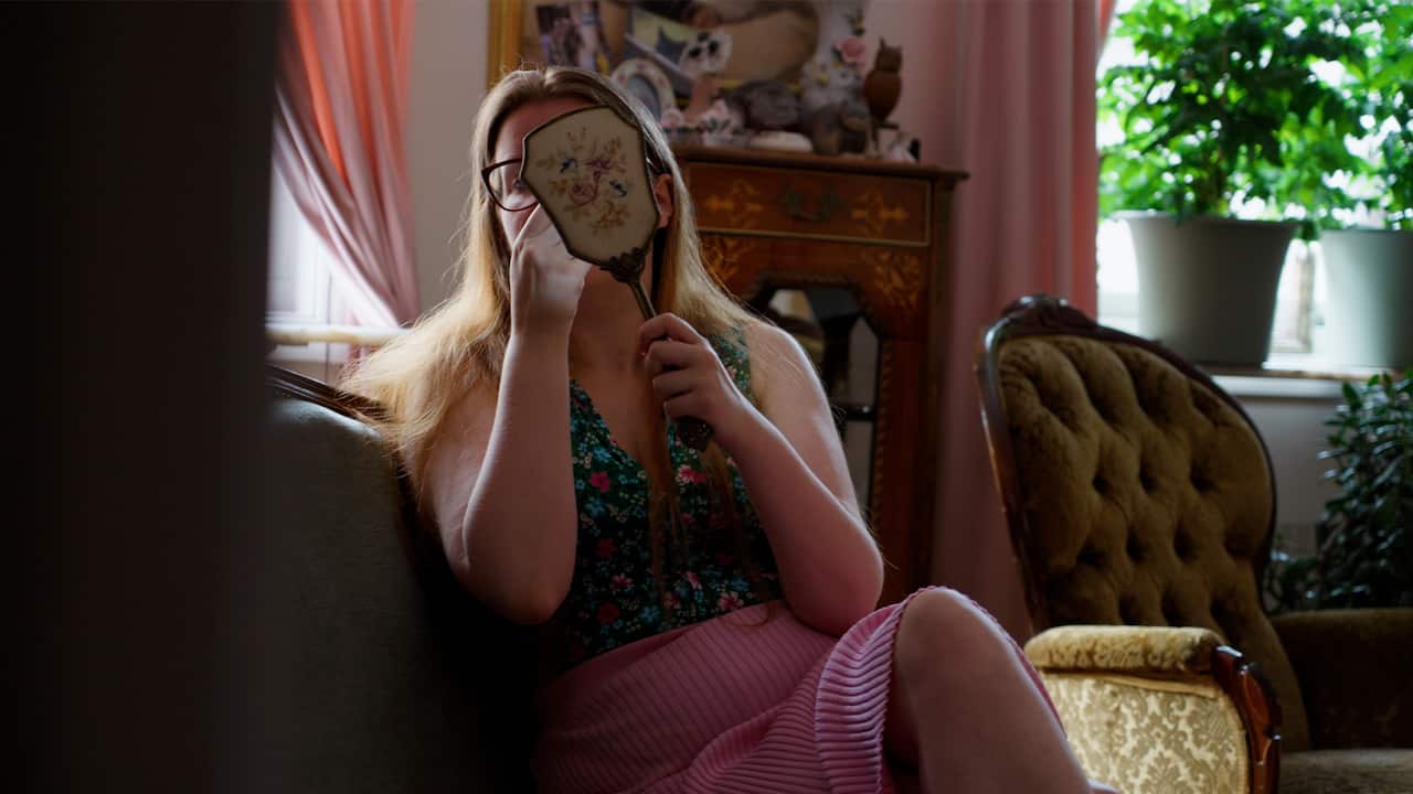 A woman with long blonde hair wearing prescription glasses, a pink skirt and a floral top is holding up a mirror to her face