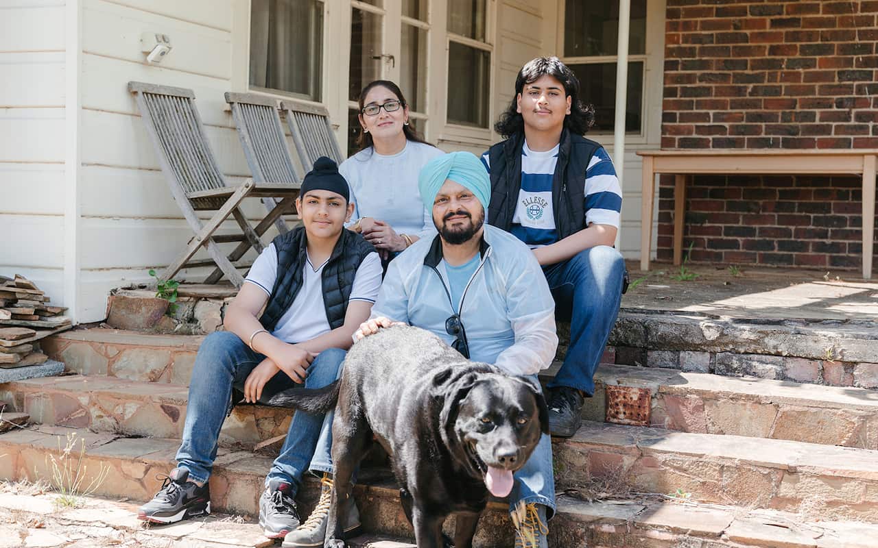A family of four sit on stairs leading off a porch. A happy looking black dog stands in front of them. 